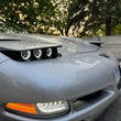 Load image into Gallery viewer, Close-up of the front C5 corvette hood and knightdrivetv headlights on silver sports car parked outdoors with trees and a concrete wall in the background.
